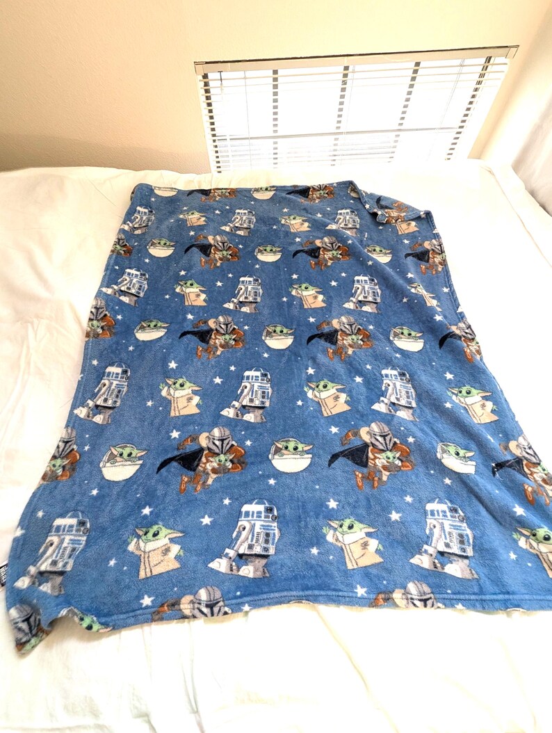 May include: A blue blanket featuring a repeating pattern of characters from a popular science fiction franchise. The design includes images of a bounty hunter, a small green alien, and a droid, along with white stars.