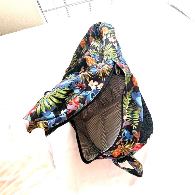May include: Black sling bag with a tropical floral and character print. The bag features a zipper closure and a mesh side pocket. The interior has a striped lining and multiple compartments. The bag is designed to be worn over one shoulder.