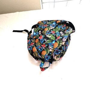 May include: A black backpack with a colorful tropical pattern featuring blue cartoon characters, pineapples, and flowers. The backpack has black straps and a handle. The design includes red, yellow, green, and blue elements.