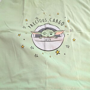 Op de afbeelding: Een lichtgroene babydeken met een afbeelding van Grogu, ook bekend als Baby Yoda, in een pod. De tekst "PRECIOUS CARGO" staat boven de pod, met gele steraccenten. De deken is opgevouwen.
