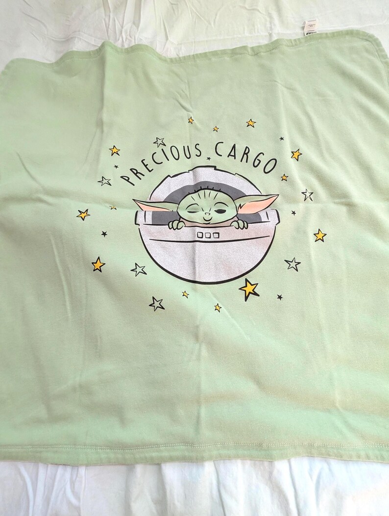 Op de afbeelding: Een lichtgroene babydeken met een afbeelding van Grogu, ook bekend als Baby Yoda, in een pod. De tekst "PRECIOUS CARGO" staat boven de pod, met gele steraccenten. De deken is opgevouwen.