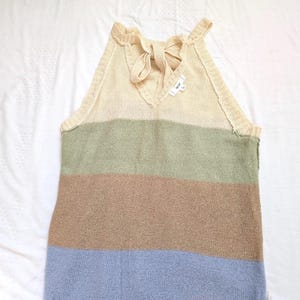 May include: A sleeveless top with a striped design in muted tones. The top has a cream-coloured upper section with a bow detail at the back, followed by horizontal stripes of green, brown, and blue. The garment is made of a knitted material.