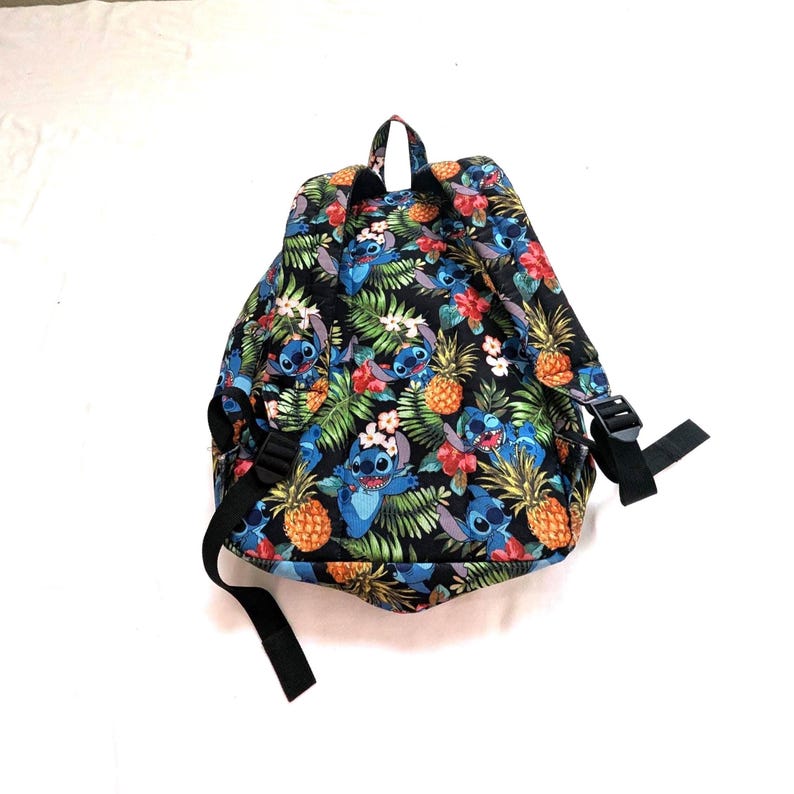 May include: Black backpack with a colorful tropical pattern featuring blue cartoon characters, pineapples, and flowers. The backpack has black straps and a top handle.