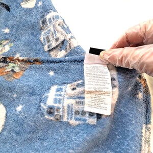 May include: A blue fleece blanket featuring a repeating pattern of white and gray illustrations of a droid and a small green character. The blanket has a soft texture and a tag with care instructions.