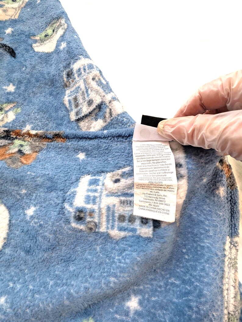 May include: A blue fleece blanket featuring a repeating pattern of white and gray illustrations of a droid and a small green character. The blanket has a soft texture and a tag with care instructions.