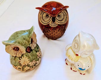 May include: Three ceramic owl figurines. One is a glossy, reddish-brown color with detailed feather patterns. Another is green with floral accents. The third is a white owl figurine with a sleeping owl on top.
