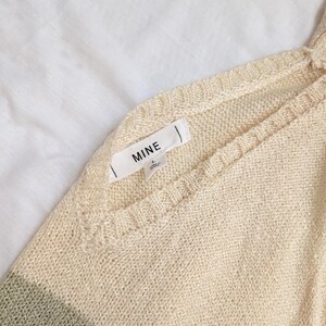 May include: A close-up shot of a cream-coloured knitted sweater with a V-neck design. The sweater has a subtle green stripe at the bottom and a tag that reads "MINE". The fabric appears soft and lightweight, suitable for layering.