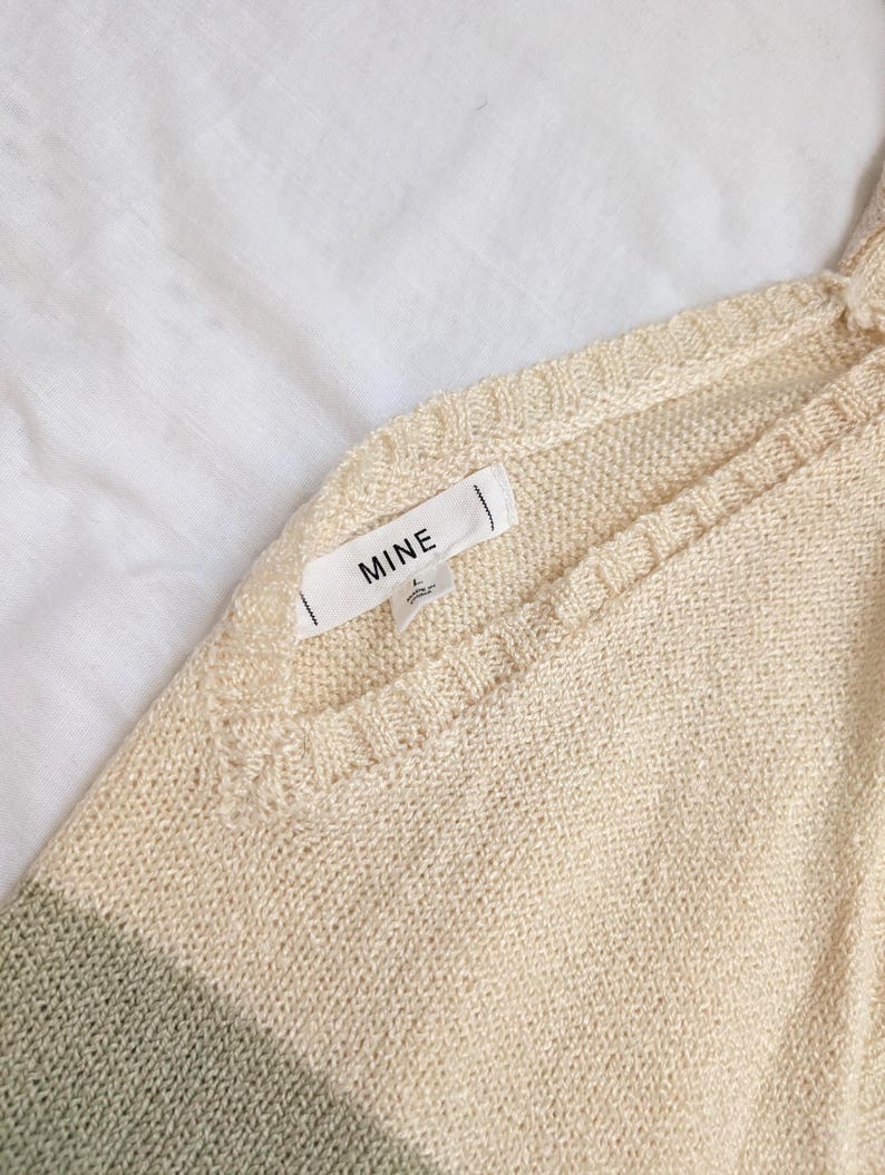 May include: A close-up shot of a cream-coloured knitted sweater with a V-neck design. The sweater has a subtle green stripe at the bottom and a tag that reads "MINE". The fabric appears soft and lightweight, suitable for layering.