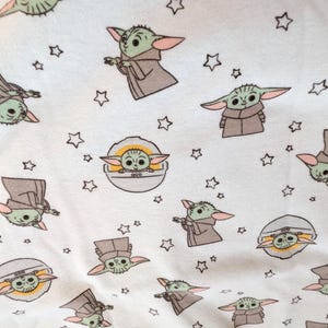 Op de afbeelding: Witte stof met een herhaald patroon van een groen alien-personage met grote oren en een bruine jurk, samen met kleine sterren. De alien wordt afgebeeld in verschillende poses en in een pod.