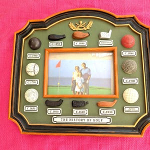 May include: A framed display with a green background, featuring golf balls and clubs from different eras, and a family photo. The frame is black and gold, with the text "THE HISTORY OF GOLF" at the bottom. The display includes various golf ball designs.