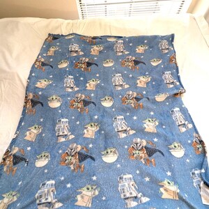 May include: A blue fleece blanket featuring repeating images of characters from a popular science fiction series. The blanket has images of a character in armor, a small green creature, and a robot. White stars are scattered across the blanket.