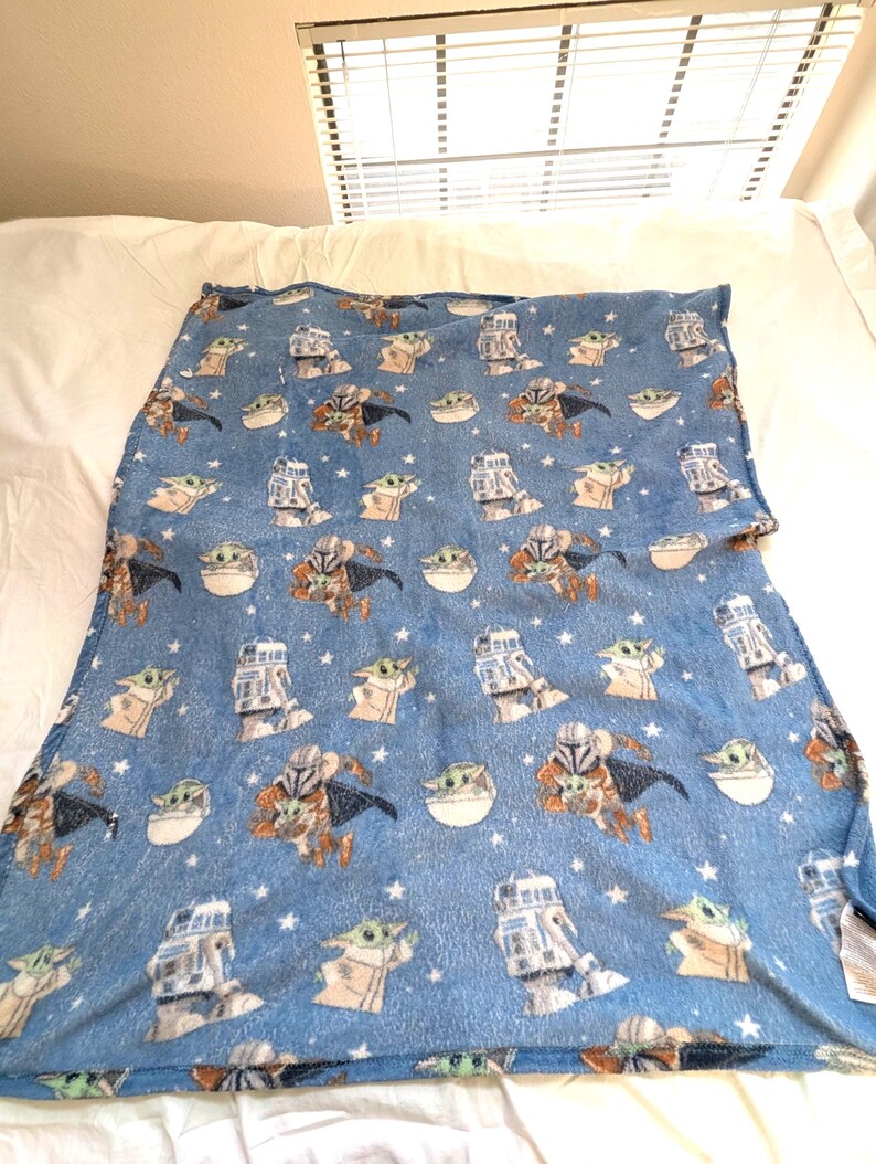 May include: A blue fleece blanket featuring repeating images of characters from a popular science fiction series. The blanket has images of a character in armor, a small green creature, and a robot. White stars are scattered across the blanket.