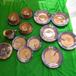 May include: A porcelain tea set featuring a metallic gold center and a purple rim decorated with black tree branches and white blossoms. The set includes a creamer, a lidded bowl, multiple cups, saucers, and plates arranged on a green fabric.