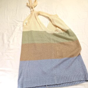 May include: A sleeveless, colour-blocked sweater top in cream, sage green, tan, and light blue. The top features a halter neck tie and a V-neckline. The bottom hem is ribbed. The sweater is made of a soft, knitted material.