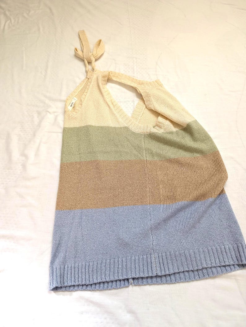 May include: A sleeveless, colour-blocked sweater top in cream, sage green, tan, and light blue. The top features a halter neck tie and a V-neckline. The bottom hem is ribbed. The sweater is made of a soft, knitted material.