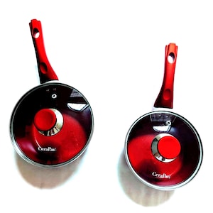 May include: Two red and silver CeraPan saucepans with glass lids and red handles. The handles are curved with a hole at the end for hanging. The pans have a red interior and a silver rim. The brand name "CeraPan" is printed on the front of the pan.
