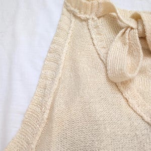 May include: A close-up shot of a cream-coloured knitted top with a V-neck and a bow detail. The top features a delicate, textured knit pattern and a subtle green stripe at the bottom. The neckline is trimmed with a decorative edge.
