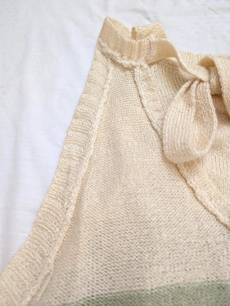 May include: A close-up shot of a cream-coloured knitted top with a V-neck and a bow detail. The top features a delicate, textured knit pattern and a subtle green stripe at the bottom. The neckline is trimmed with a decorative edge.