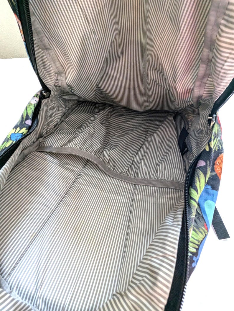 May include: Open backpack interior with a gray and white striped lining. The backpack's exterior features a dark background with colorful floral and bird patterns. The zipper is black.