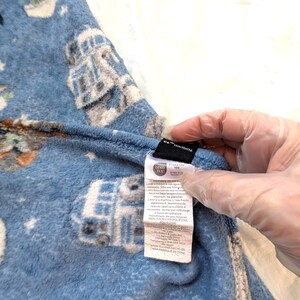 May include: Blue fleece pajama pants with a Star Wars-themed pattern featuring characters and vehicles. The pants have a tag with care instructions and material information. The fabric is a soft, plush material.