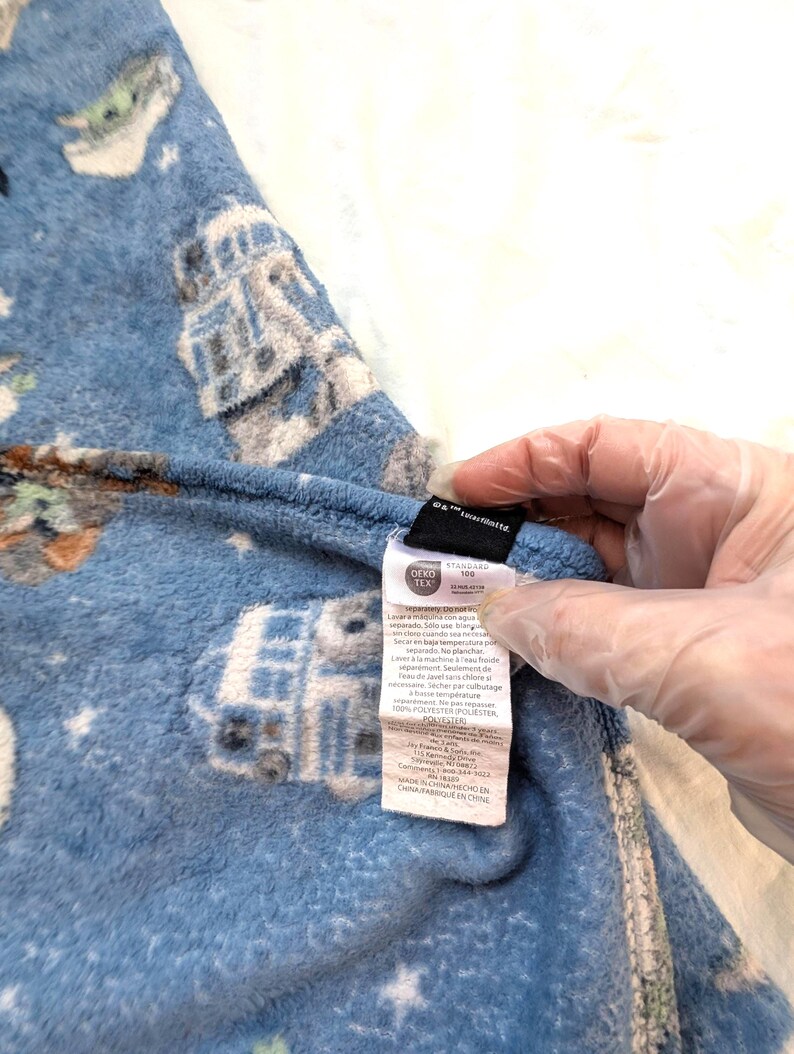May include: Blue fleece pajama pants with a Star Wars-themed pattern featuring characters and vehicles. The pants have a tag with care instructions and material information. The fabric is a soft, plush material.