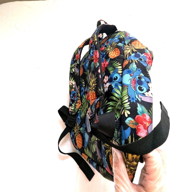 May include: Black backpack with a tropical print featuring pineapples, flowers, and the character Stitch. The backpack has black straps and a side pocket. The design includes blue, red, yellow, and green elements.