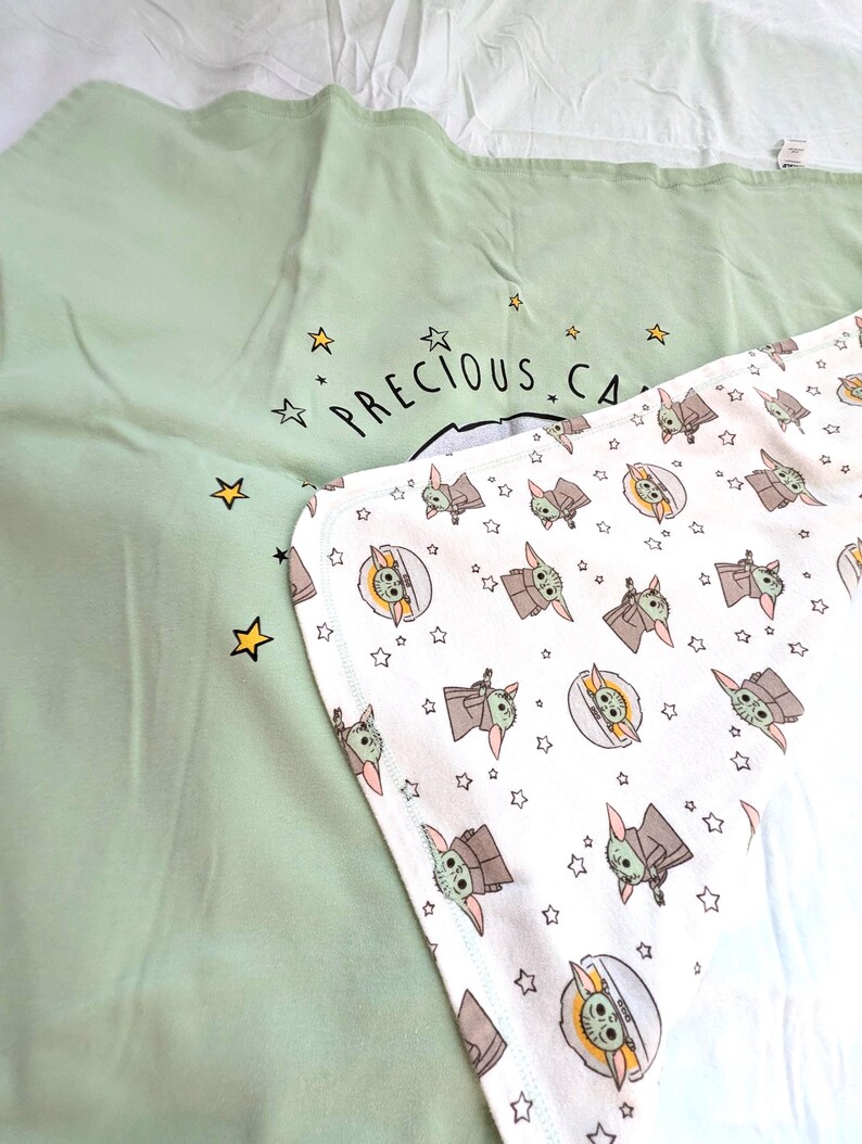 Op de afbeelding: Twee babydekens. E&eacute;n is lichtgroen met de tekst "PRECIOUS CA..." en gele sterren. De andere is wit met een patroon van een stripfiguur en sterren. De dekens zijn op een wit oppervlak gevouwen.