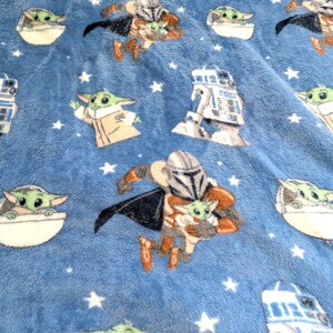 May include: A blue plush blanket featuring repeating images of The Mandalorian, Grogu, and R2-D2. The blanket has a soft texture and is decorated with white stars. The characters are in various poses.
