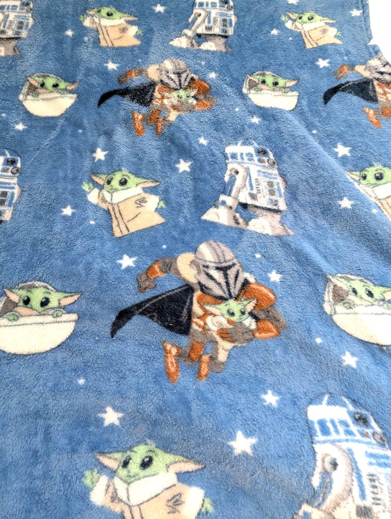 May include: A blue plush blanket featuring repeating images of The Mandalorian, Grogu, and R2-D2. The blanket has a soft texture and is decorated with white stars. The characters are in various poses.