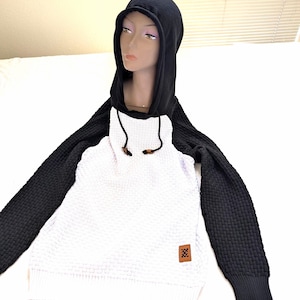 May include: A black and white hooded sweater with a textured knit pattern. The sleeves and hood are black, while the body is white. The sweater features a drawstring and a small brown patch with a logo.