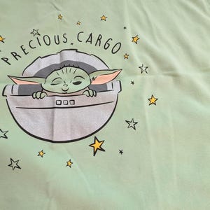 Op de afbeelding: Lichtgroene stof met een afbeelding van een personage uit een populaire sciencefiction-serie. Het personage zit in een zilveren pod met de woorden "PRECIOUS CARGO" erboven. Gele sterren zijn verspreid rond de afbeelding.