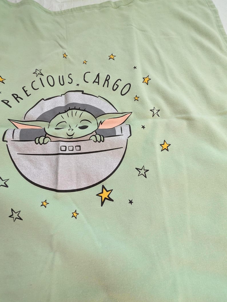 Op de afbeelding: Lichtgroene stof met een afbeelding van een personage uit een populaire sciencefiction-serie. Het personage zit in een zilveren pod met de woorden "PRECIOUS CARGO" erboven. Gele sterren zijn verspreid rond de afbeelding.