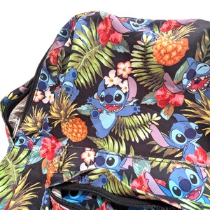 May include: A black bag with a colorful tropical pattern featuring the character Stitch from Lilo & Stitch. The design includes Stitch, pineapples, red hibiscus flowers, and green palm leaves. The bag has a zipper closure.