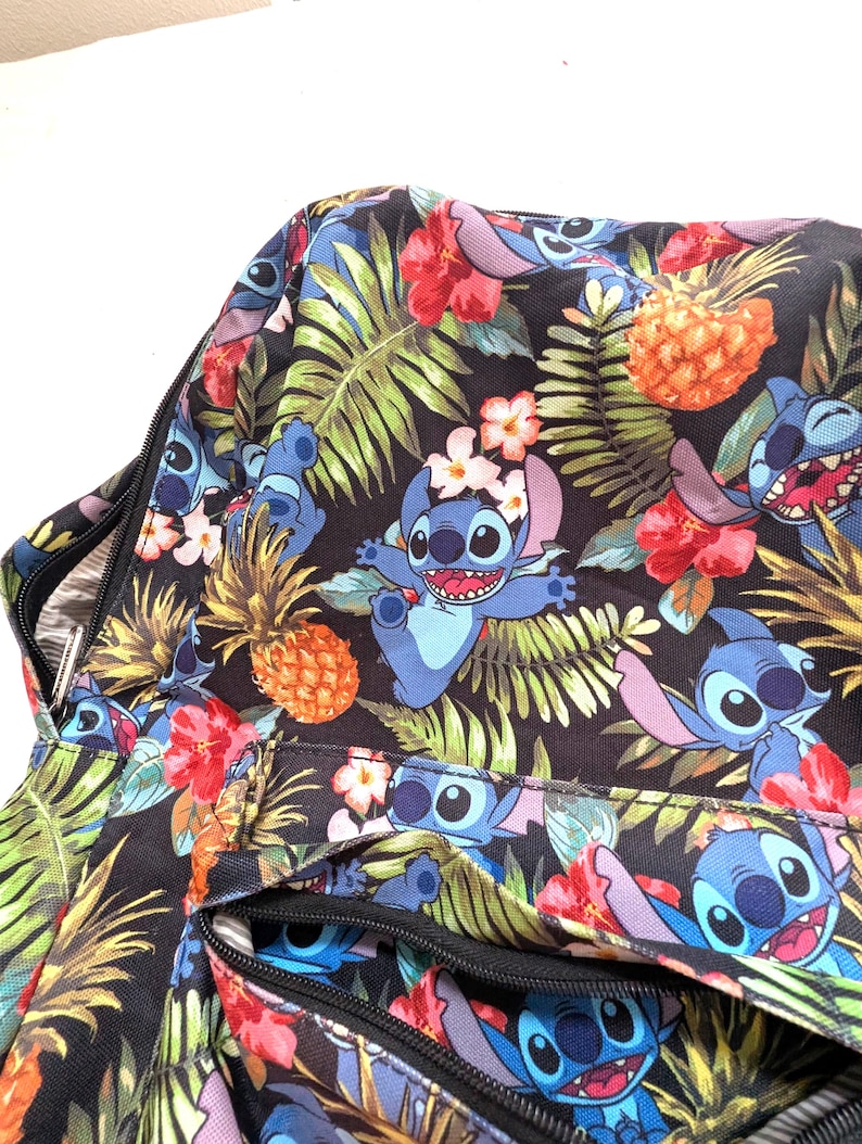 May include: A black bag with a colorful tropical pattern featuring the character Stitch from Lilo & Stitch. The design includes Stitch, pineapples, red hibiscus flowers, and green palm leaves. The bag has a zipper closure.