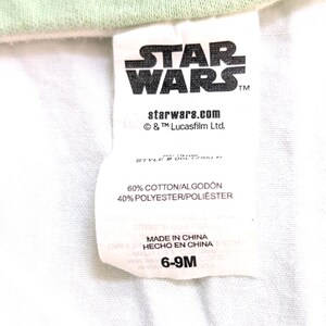 Op de afbeelding: Een wit label met het Star Wars-logo in zwart, het websiteadres starwars.com en de tekst "6-9M". Het label vermeldt ook dat de stof bestaat uit 60% katoen en 40% polyester.