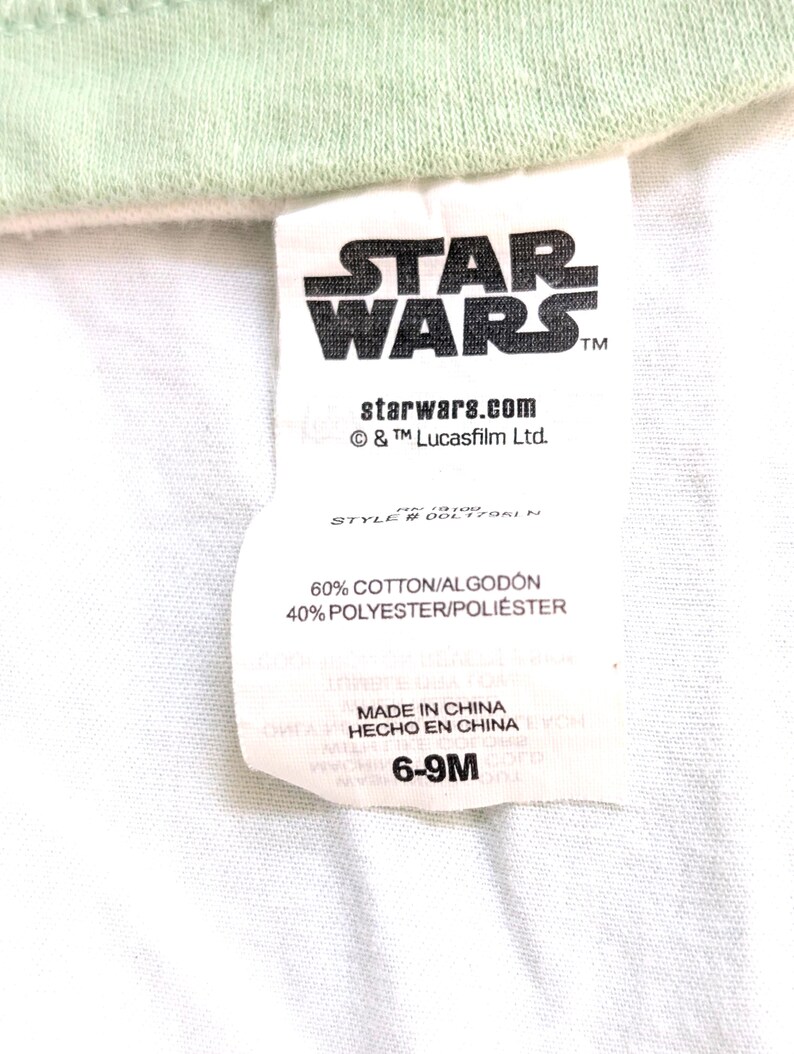 Op de afbeelding: Een wit label met het Star Wars-logo in zwart, het websiteadres starwars.com en de tekst "6-9M". Het label vermeldt ook dat de stof bestaat uit 60% katoen en 40% polyester.