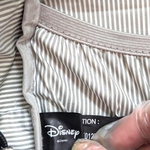 May include: Close-up of a black Disney label with the Disney logo and text. The label is inside a bag with a gray and white striped lining. The bag has a gray elastic band.