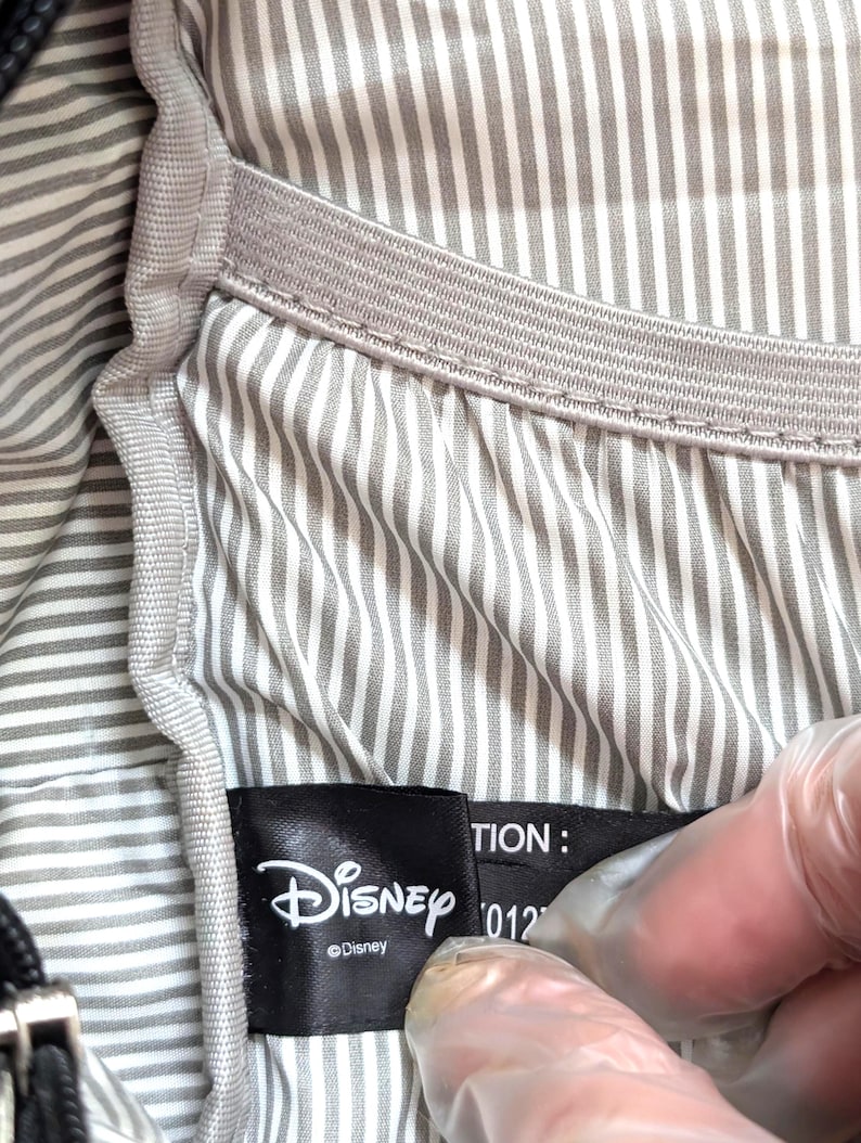 May include: Close-up of a black Disney label with the Disney logo and text. The label is inside a bag with a gray and white striped lining. The bag has a gray elastic band.