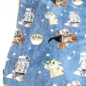 May include: A blue plush blanket featuring repeating images of a green alien character, a droid, and a character in armor. White stars are scattered across the blanket. The blanket is made of a soft, fuzzy material.