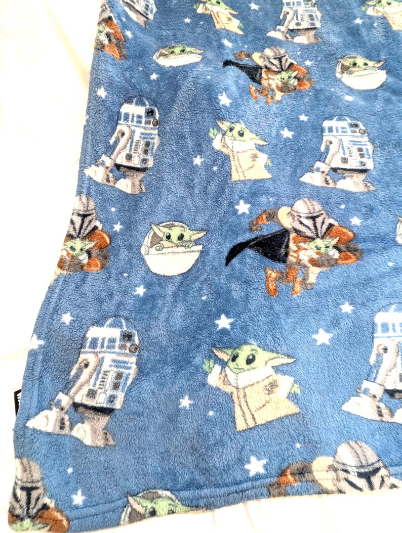 May include: A blue plush blanket featuring repeating images of a green alien character, a droid, and a character in armor. White stars are scattered across the blanket. The blanket is made of a soft, fuzzy material.