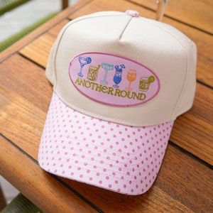 May include: A beige baseball cap with a pink polka dot brim. The front features an oval patch with embroidered colorful cocktail glasses and the text "ANOTHER ROUND" in gold lettering. The cap has a structured crown and a button on top.