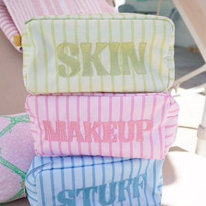May include: Three striped cosmetic bags stacked on top of each other. The top bag is light green with the word "SKIN" embroidered in a darker green. The middle bag is pink with "MAKEUP" embroidered in pink. The bottom bag is blue with "STUFF" embroidered in blue.