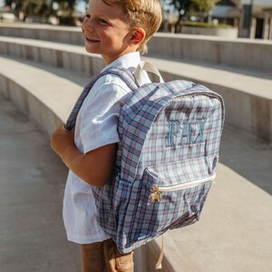 May include: A blue, white, and red plaid backpack featuring the name PAX embroidered in blue thread on the front pocket. The backpack has a white zipper, white shoulder straps, and a gold-toned zipper pull on the front compartment.