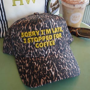 May include: A leopard print baseball cap with yellow embroidered text that reads SORRY I'M LATE, I STOPPED FOR COFFEE. The cap is positioned in front of a white canvas tote bag with black and white striped handles and the green embroidered letters HW.