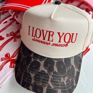 May include: Three baseball caps with different designs. The first cap is white with red trim and says "Leave Him On Read". The second cap is white with a leopard print brim and says "I Love You soooo much". The third cap is pink with white text that says "XOXO".