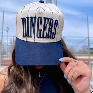 May include: A baseball-style cap with a navy blue brim and a beige crown with vertical pinstripes. The word "DINGERS" is embroidered in navy blue across the front. The cap has a button on top.