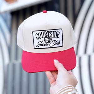 May include: A cream-colored Courtside Social Club baseball cap with a red brim and top button. The cap features a black and white rectangular patch with the club's name and a basketball graphic.