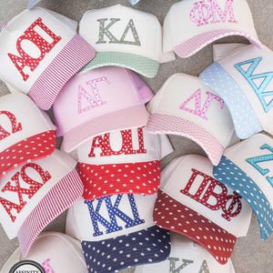 May include: A collection of white trucker hats featuring various Greek letter combinations in colorful patterns. Each hat has a structured front panel with embroidered letters and a matching patterned brim, including polka dots and stripes in red, blue, and pink.