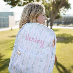 May include: A backpack featuring a white fabric with vertical light pink stripes and a repeating floral pattern of small pink roses and green leaves. The name Henley is embroidered in pink script on the front, with rose gold zipper pulls.
