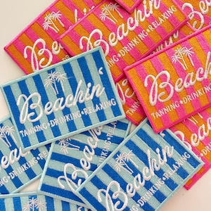 May include: A collection of rectangular embroidered patches featuring vertical stripes in either pink and orange or light and dark blue. Each patch displays the white embroidered text Beachin, a palm tree icon, and the words TANNING DRINKING RELAXING.
